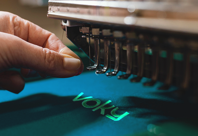 Ljusgr&ouml;n Voky-logotyp broderad p&aring; bl&aring;tt tyg.