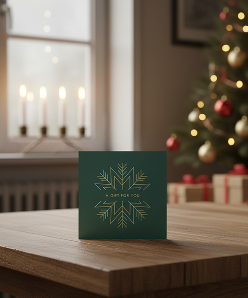 Ett gr&ouml;nt presentkort med gyllene sn&ouml;flinga och texten "A GIFT FOR YOU" p&aring; ett bord, med adventsljusstake och julgran i bakgrunden.